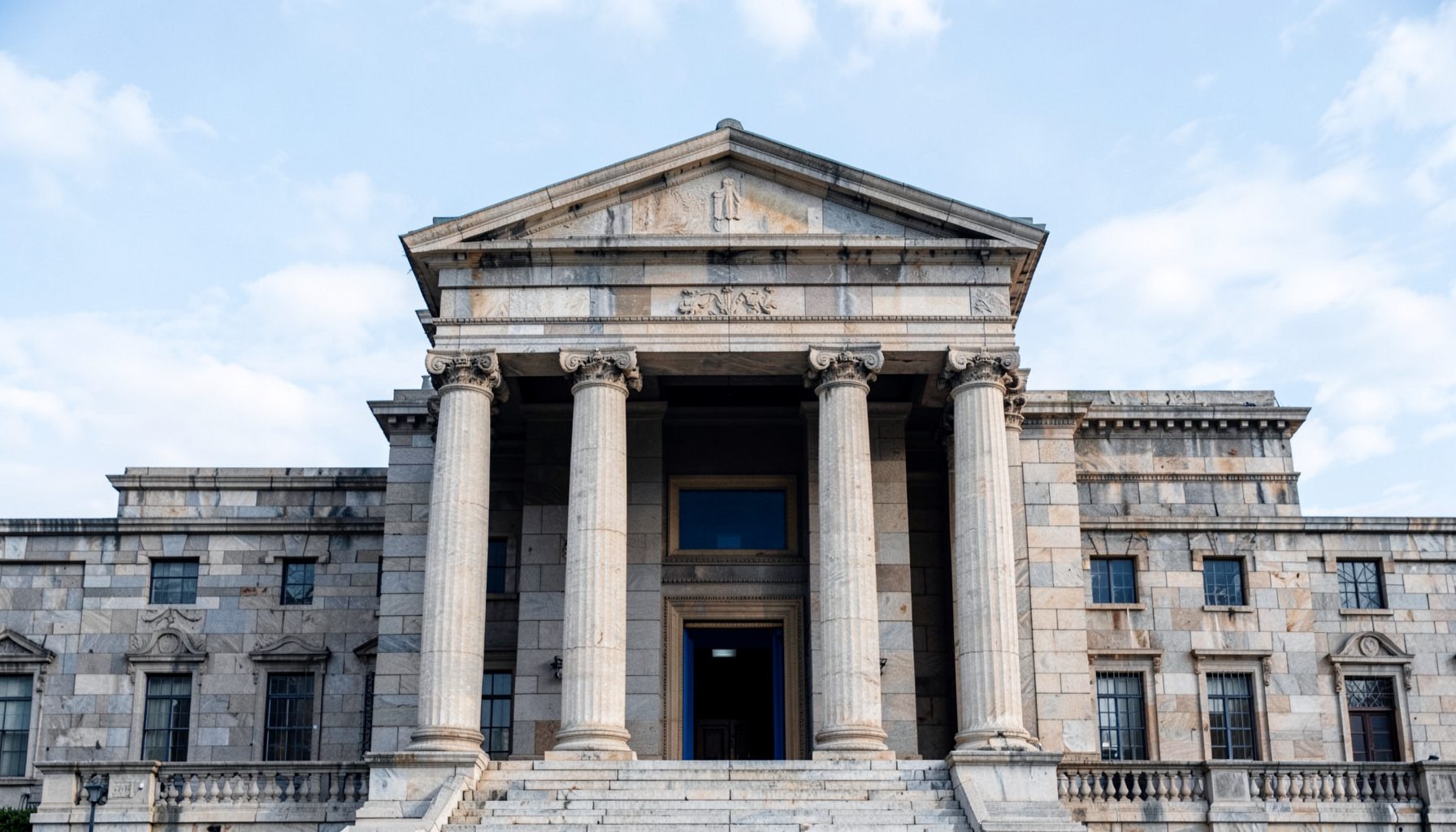 Symmetrical facade of a stone civic building with tall columns, set against a pale blue sky with soft white clouds, conveying a calm and authoritative tone in navy and warm gray tones