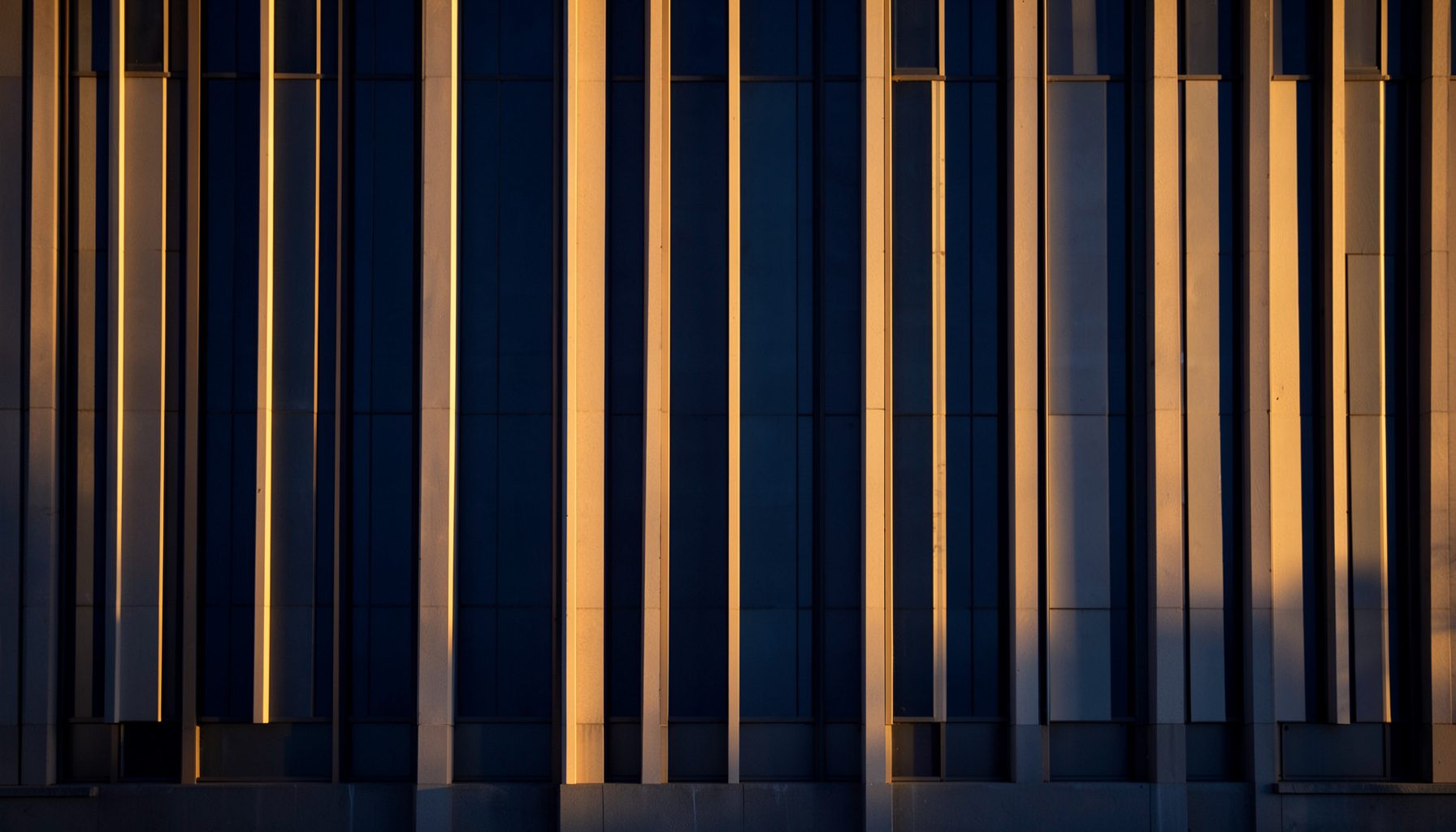 Tall vertical lines of a civic building facade at golden hour, deep navy and warm amber tones, calm and orderly atmosphere